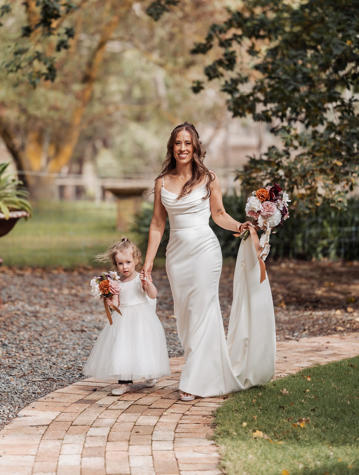 the lane vineyard wedding - caitlyn & craig 93 Bride and flower girl walking in garden, holding bouquets, with sunlight filtering through trees.