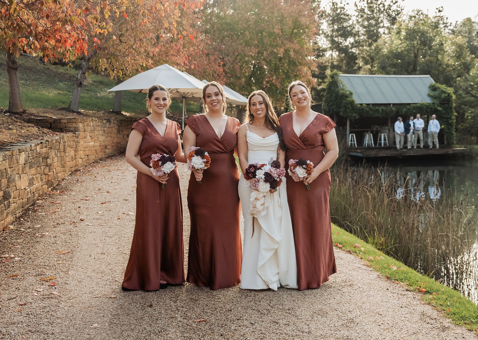 the lane vineyard wedding - caitlyn & craig 115 Bridal party by pond with autumn foliage, featuring bride in white and rust-colored bridesmaid dresses.