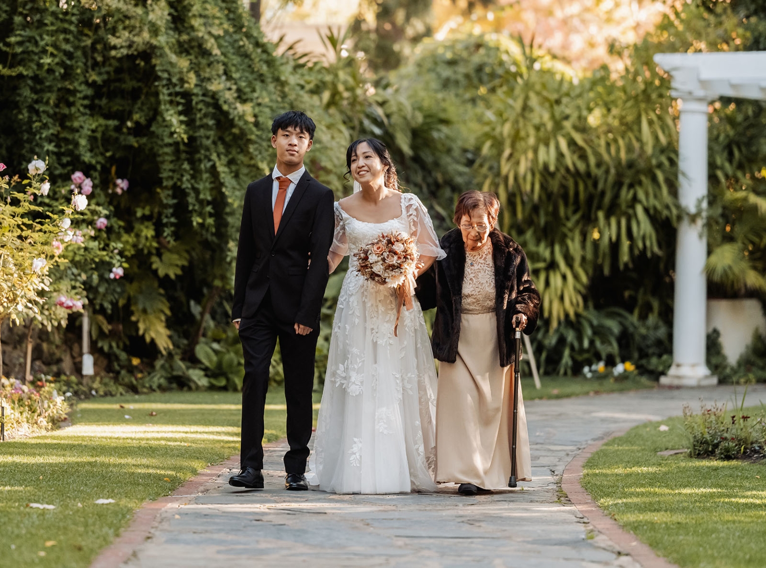 adelaide hills wedding at the manor - jo & dan 19 Bride walks garden wedding path with family, bouquet, and elderly relative.
