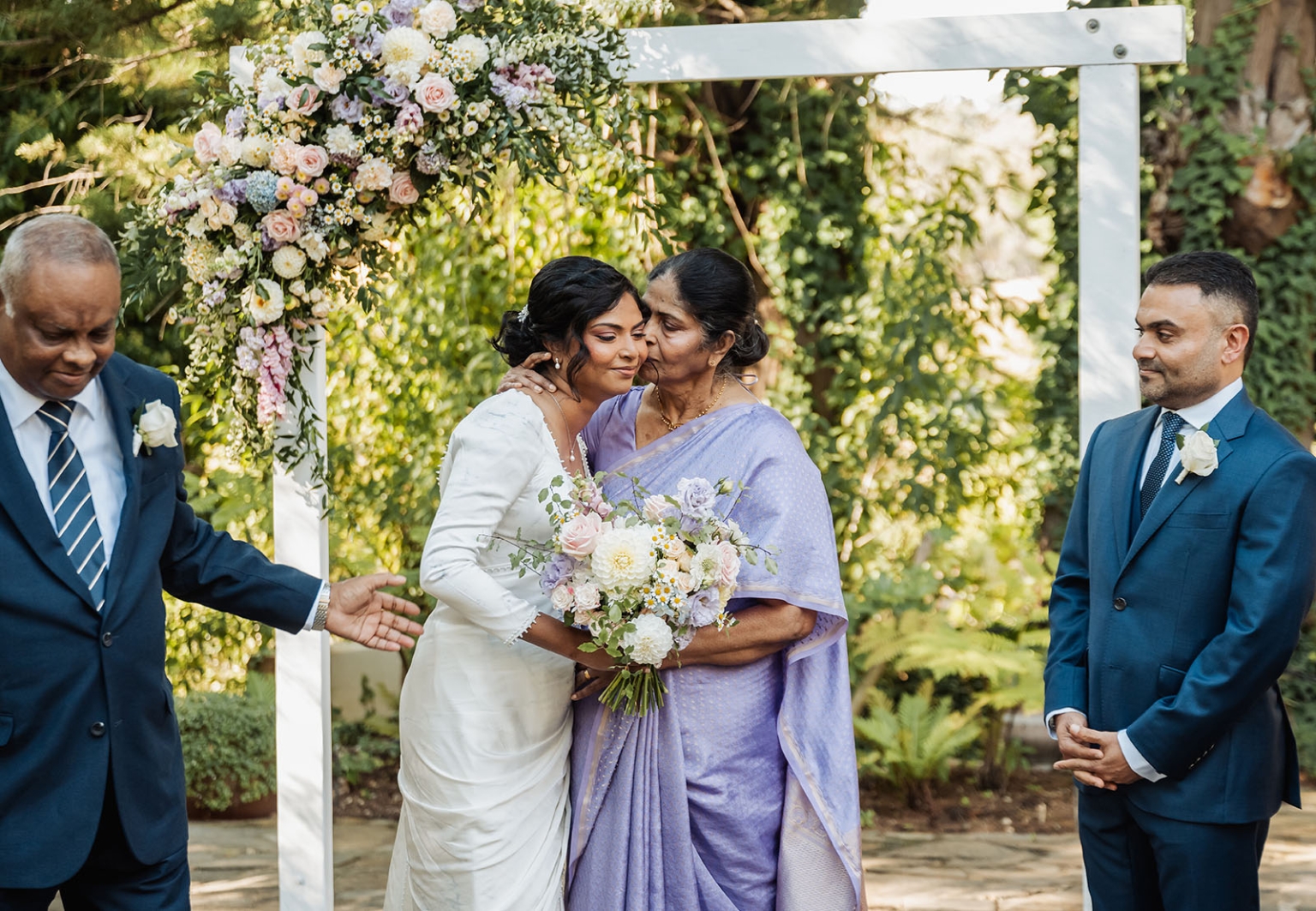 manor wedding in basket range - methmini & aravinda 135 Outdoor wedding scene with bride, affectionate family, floral arch, and lush greenery.
