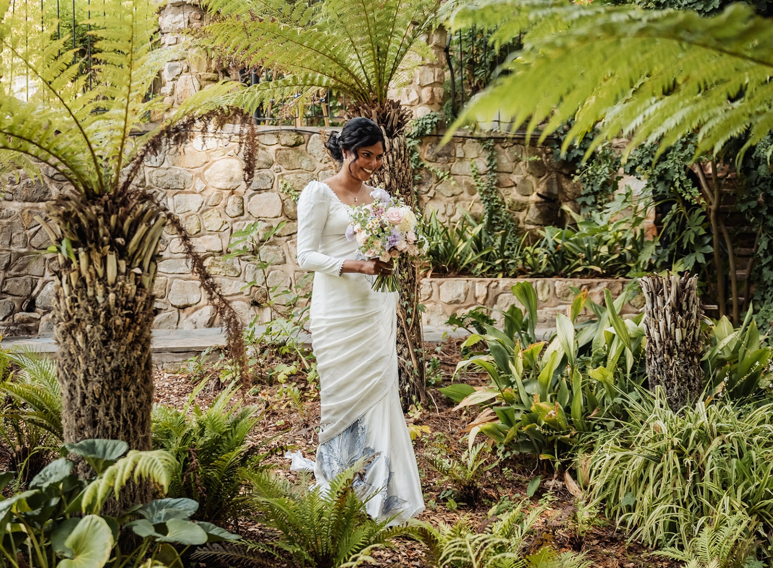 manor wedding in basket range - methmini & aravinda 122 Bride in lush garden, white dress, bouquet, stone wall backdrop, serene wedding scene.