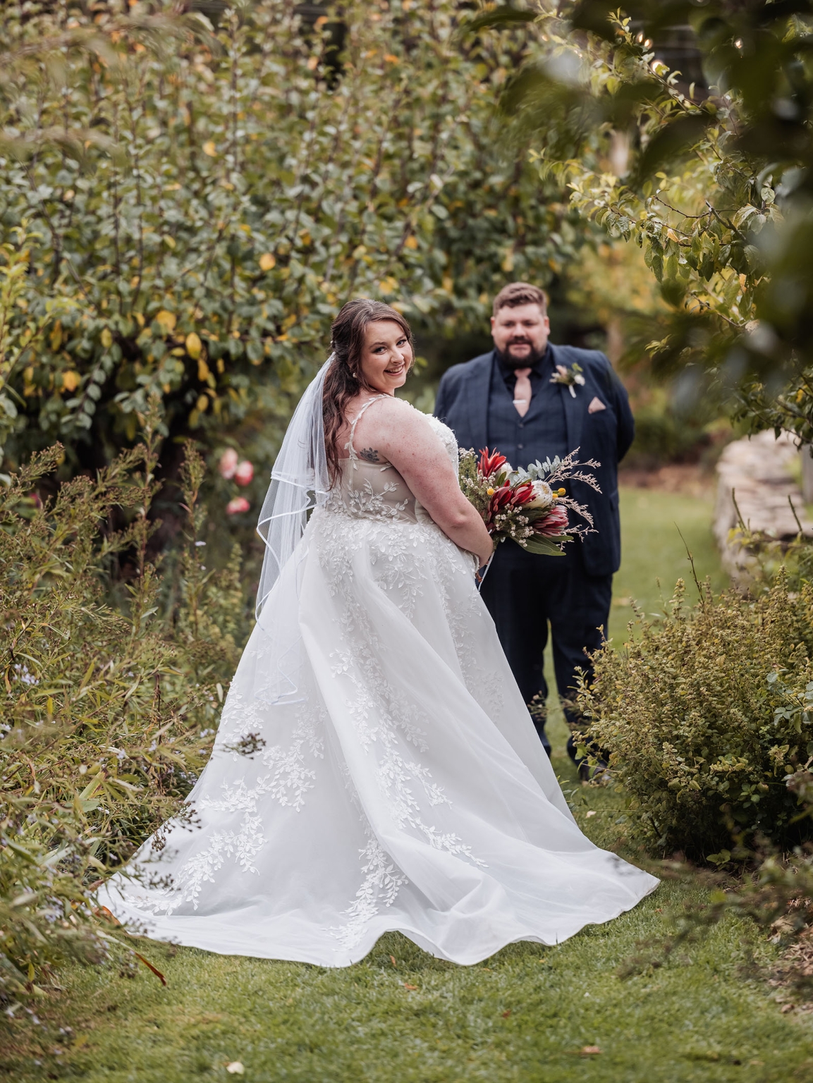 comida wedding - georgia & james 35 Bride in elegant lace gown holding bouquet, garden wedding backdrop, groom in dark suit.