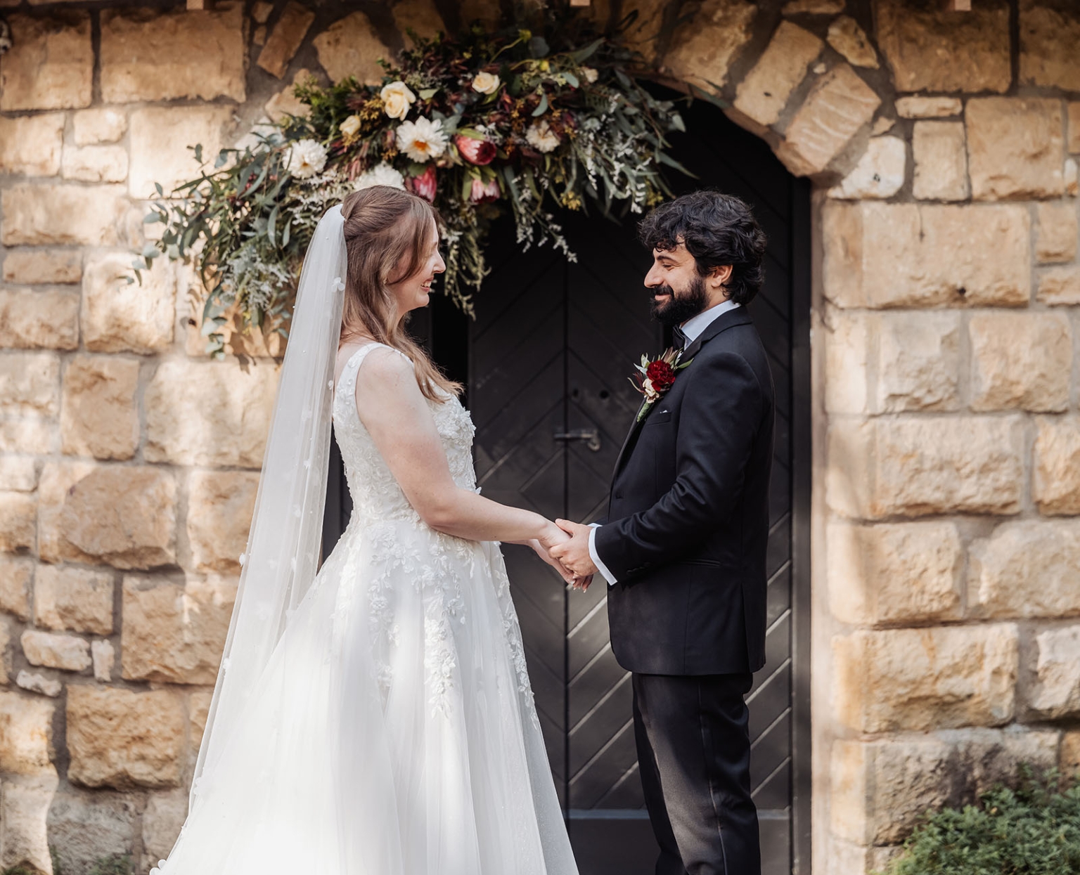 beautiful manor basket range wedding - sophie & theodore 103 Bride and groom embrace in front of rustic stone building with floral arch.