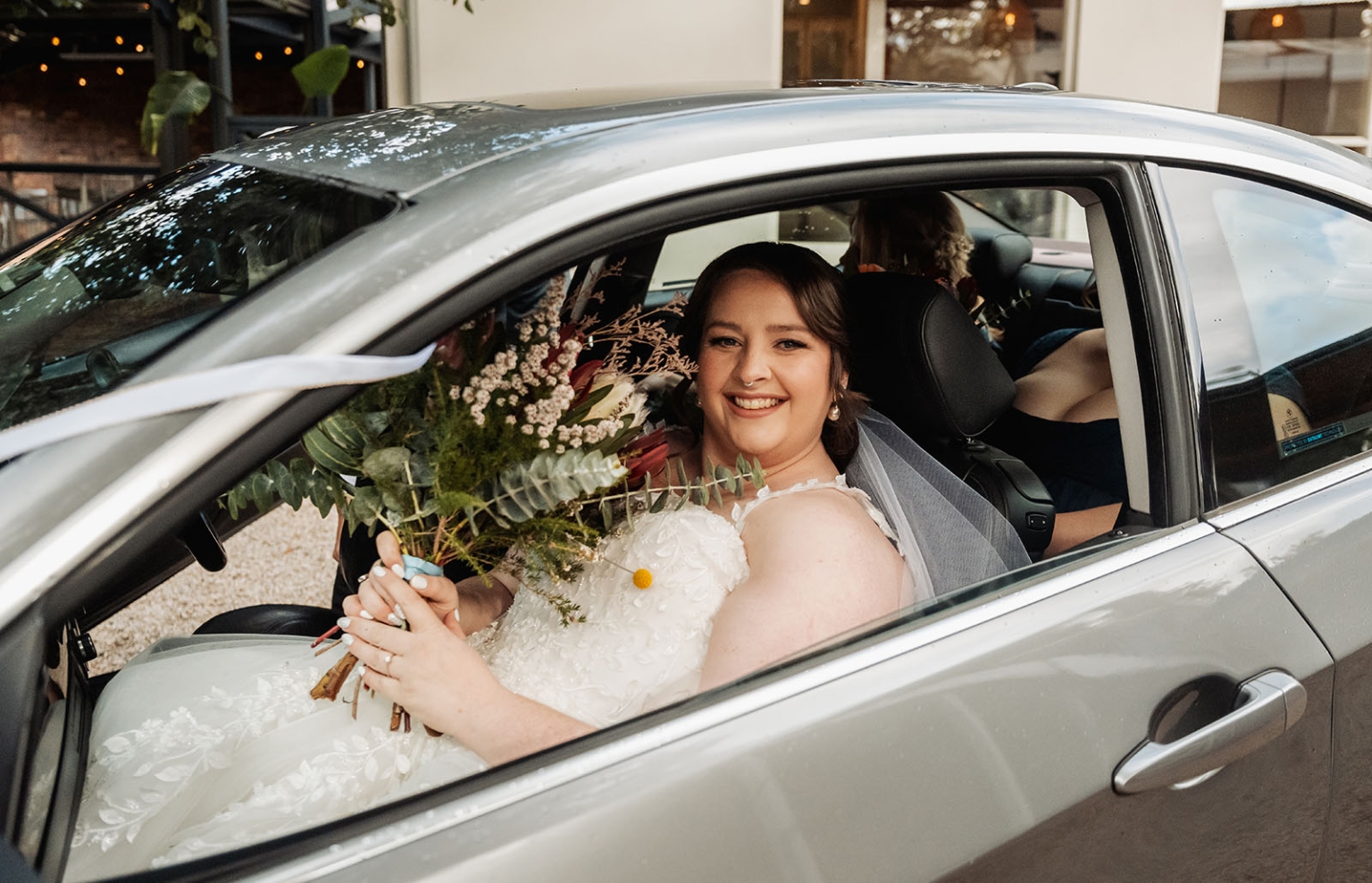 comida wedding - georgia & james 8 Joyful bride in car, white dress, holding vibrant bouquet, smiling, window view.
