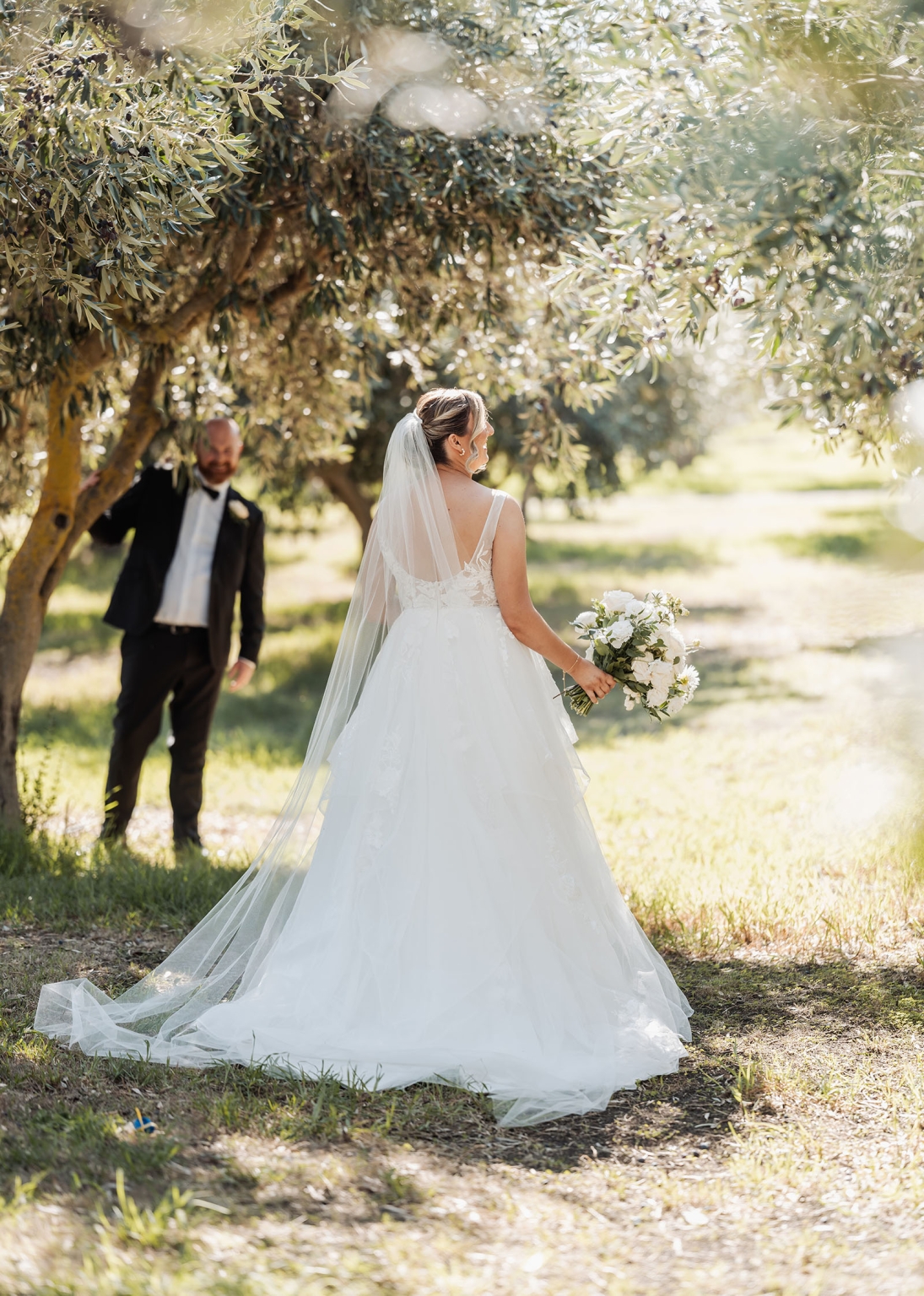 lloyd brothers wedding - dajana & tom 38 Bride in lace gown and veil under trees, holding white bouquet, groom in background.