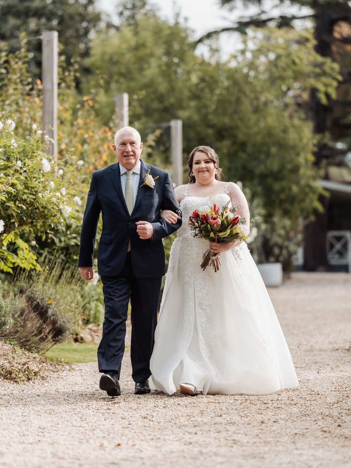 comida wedding - georgia & james 12 Bride in lace dress walking with father outdoors, holding vibrant bouquet, during wedding ceremony.