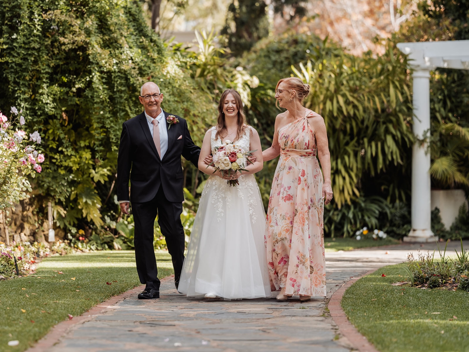 beautiful manor basket range wedding - sophie & theodore 99 Bride walks with parents in a garden wedding, wearing a lace gown with pastel flowers.