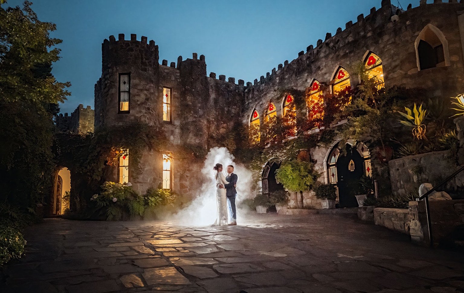 manor wedding in basket range - methmini & aravinda 161 Romantic couple embraces in a medieval castle courtyard at twilight, surrounded by stained glass light.
