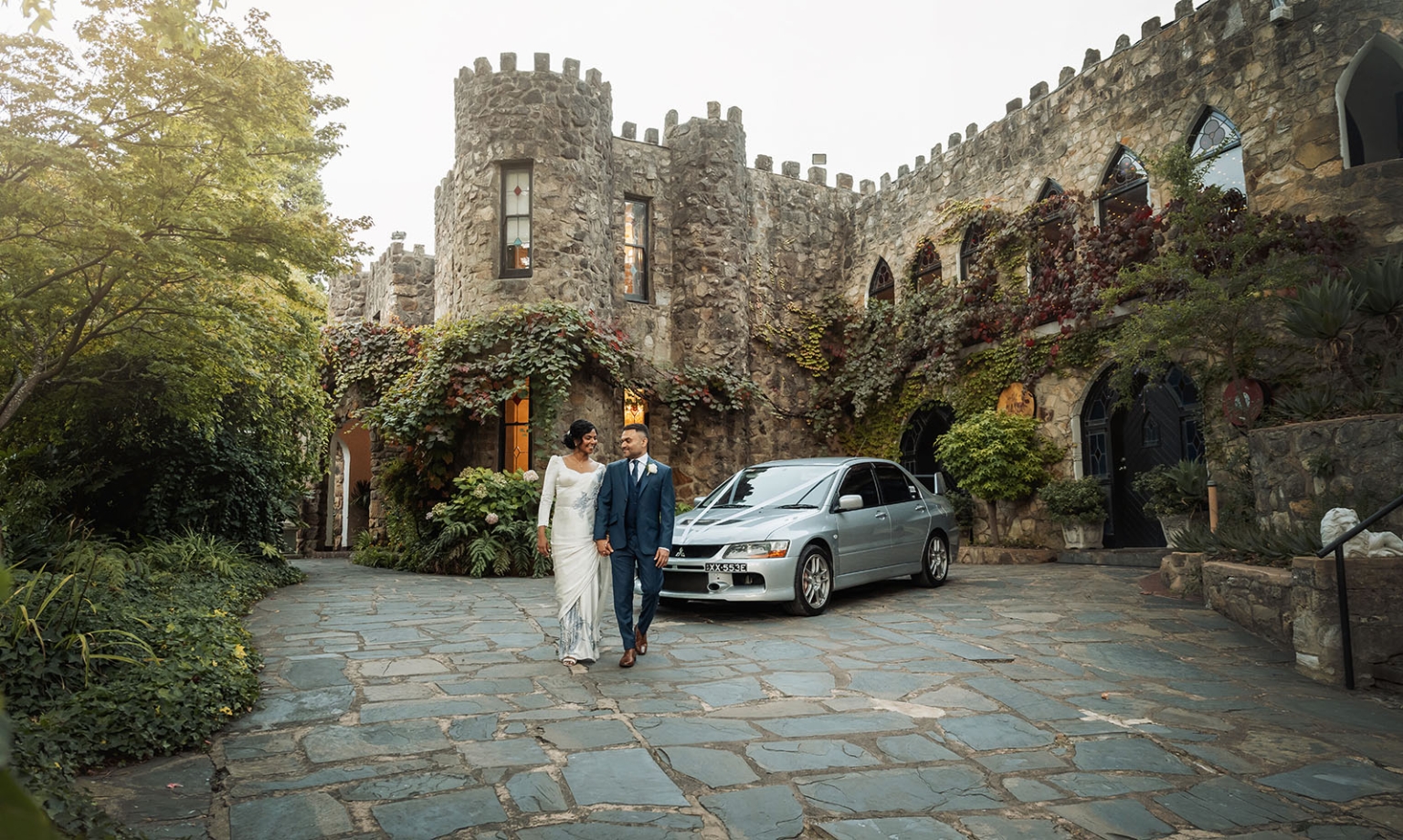 manor wedding in basket range - methmini & aravinda 158 Elegant couple in wedding attire outside a historic castle with silver car and lush greenery.