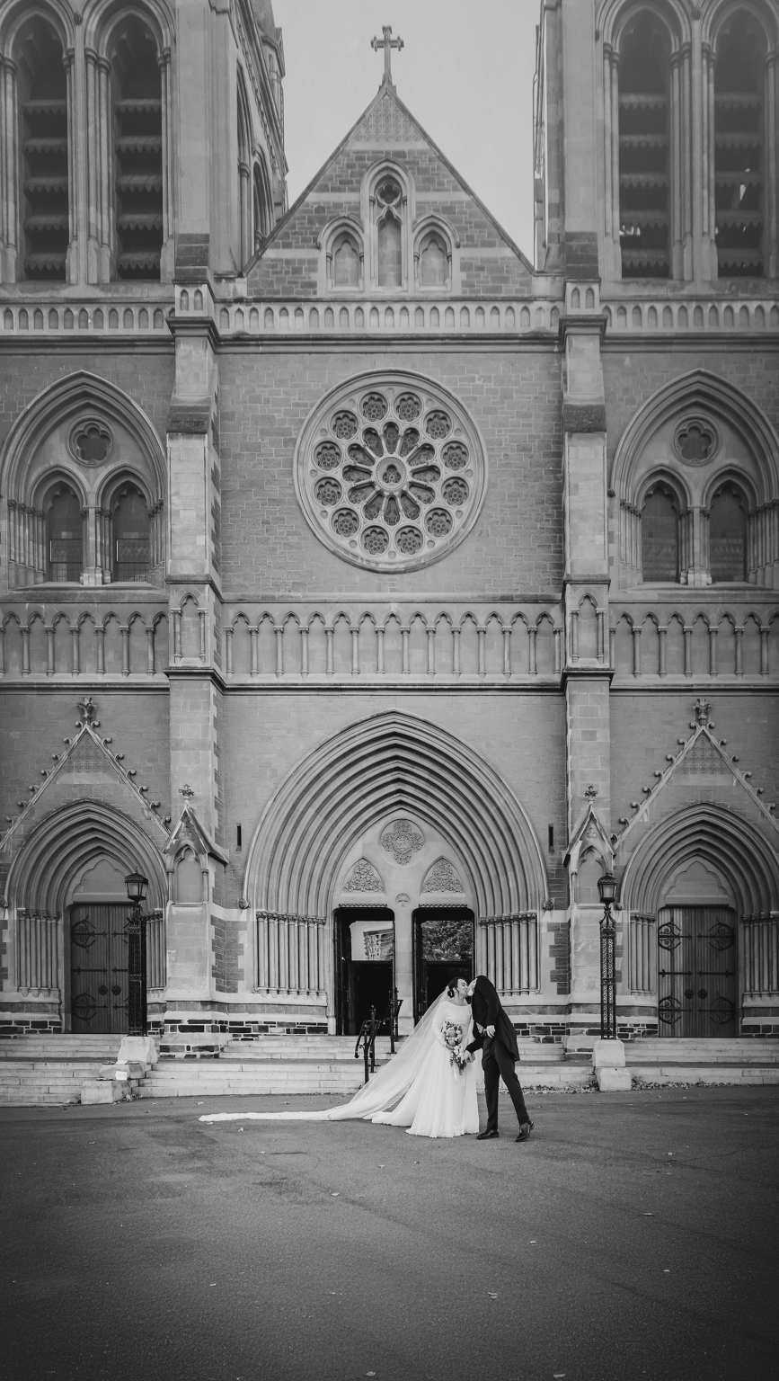 adelaide's st peter's cathedral wedding - irish & nick 33 Bride and groom kissing before a Gothic cathedral in a timeless black-and-white wedding portrait.