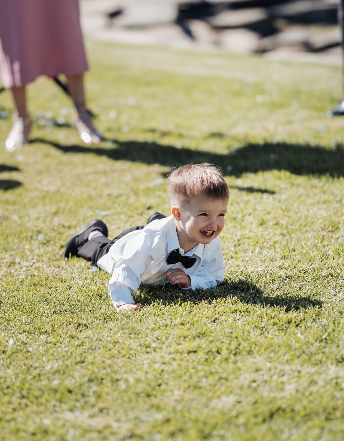 lloyd brothers wedding - dajana & tom 18 Young boy in formal attire joyfully crawls on grass at a sunny garden event.