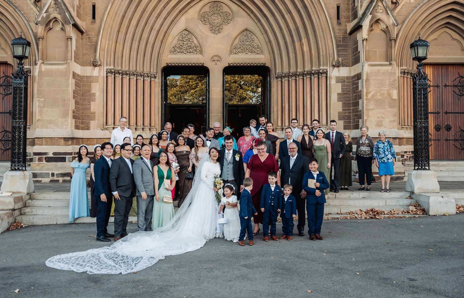 adelaide's st peter's cathedral wedding - irish & nick 31 Wedding party portrait outside historic stone church with bride, groom, family, and guests.
