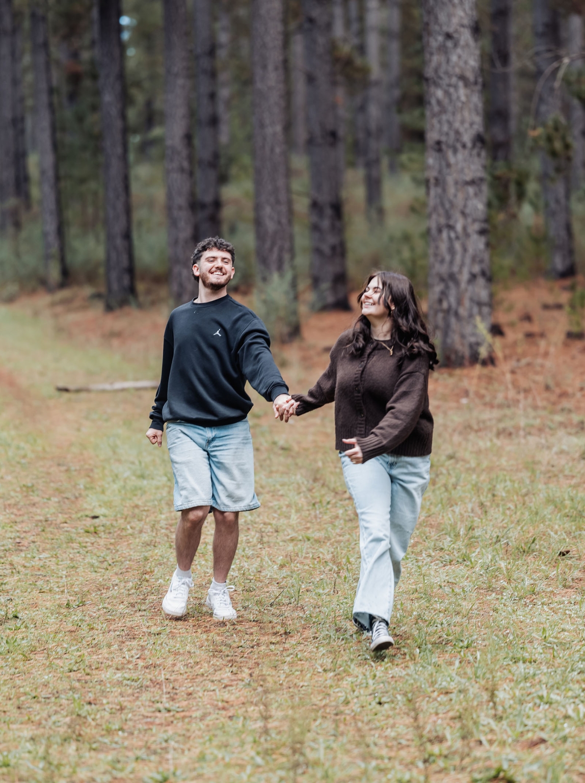 cabn at kuitpo forest proposal - tamsyn & blake 16 Couple joyfully walking in a sunlit forest path, surrounded by tall trees and natural beauty.