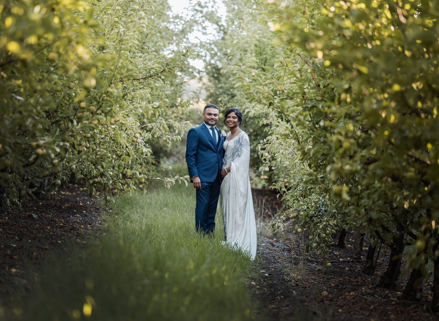 manor wedding in basket range - methmini & aravinda 153 Couple in formal attire standing lovingly in a sunlit orchard path, lush greenery around them.