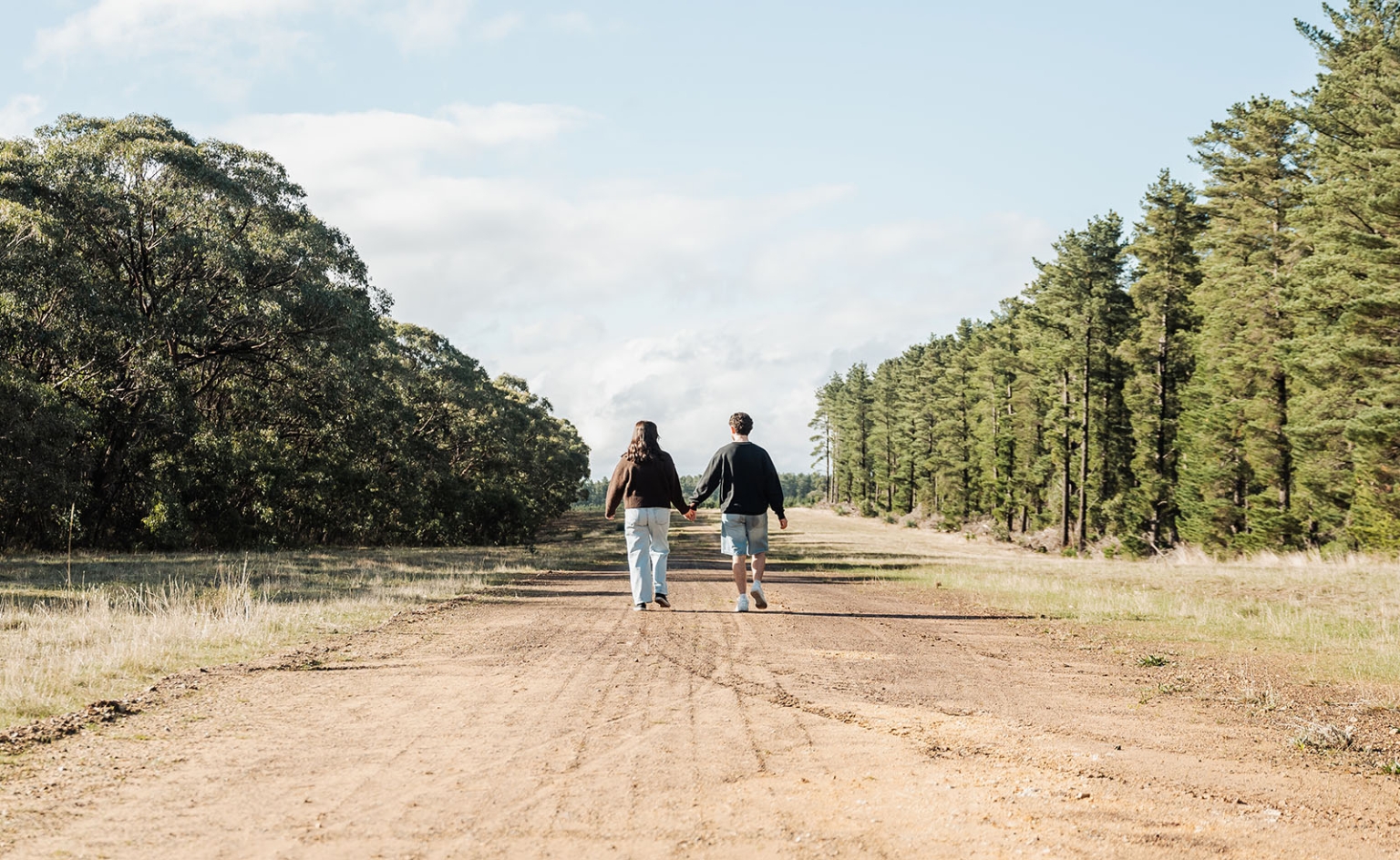 cabn at kuitpo forest proposal - tamsyn & blake 33 Couple walking hand in hand on a forest road under a serene sky.