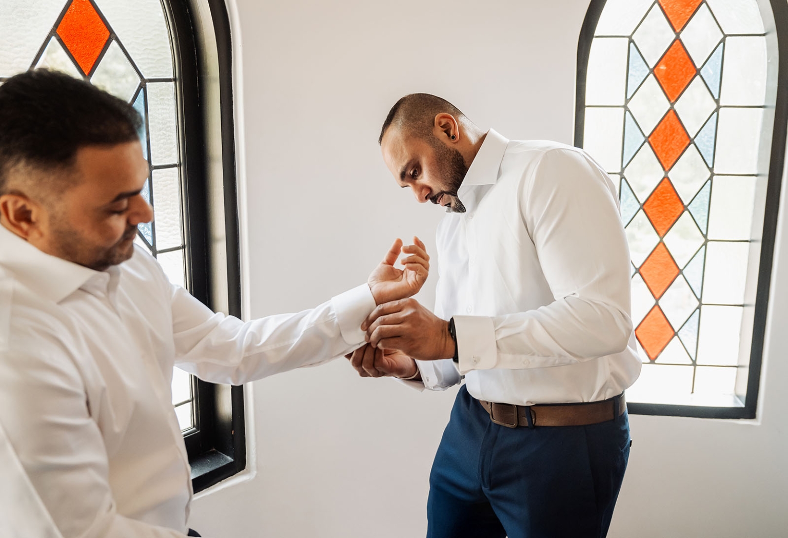 manor wedding in basket range - methmini & aravinda 97 Two men dressing with focus and camaraderie, adjusting cufflinks near stained glass windows.