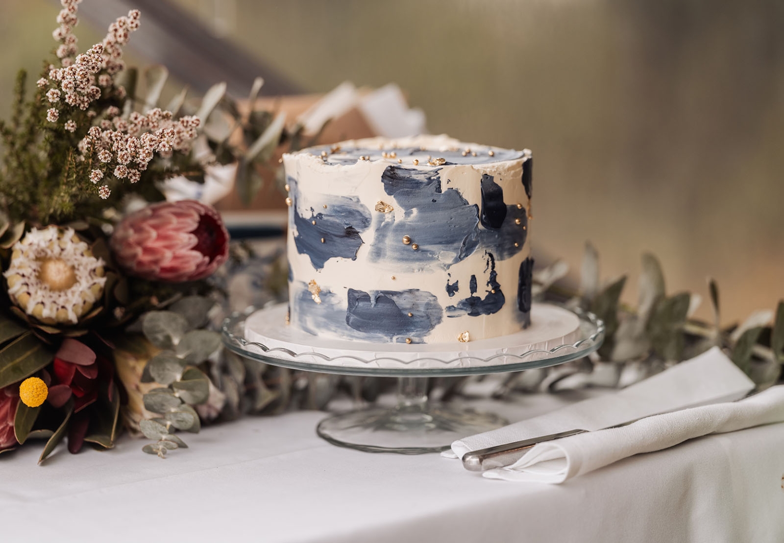 comida wedding - georgia & james 44 Elegant cake with blue and gold design, protea flowers, glass stand, for luxury celebration.