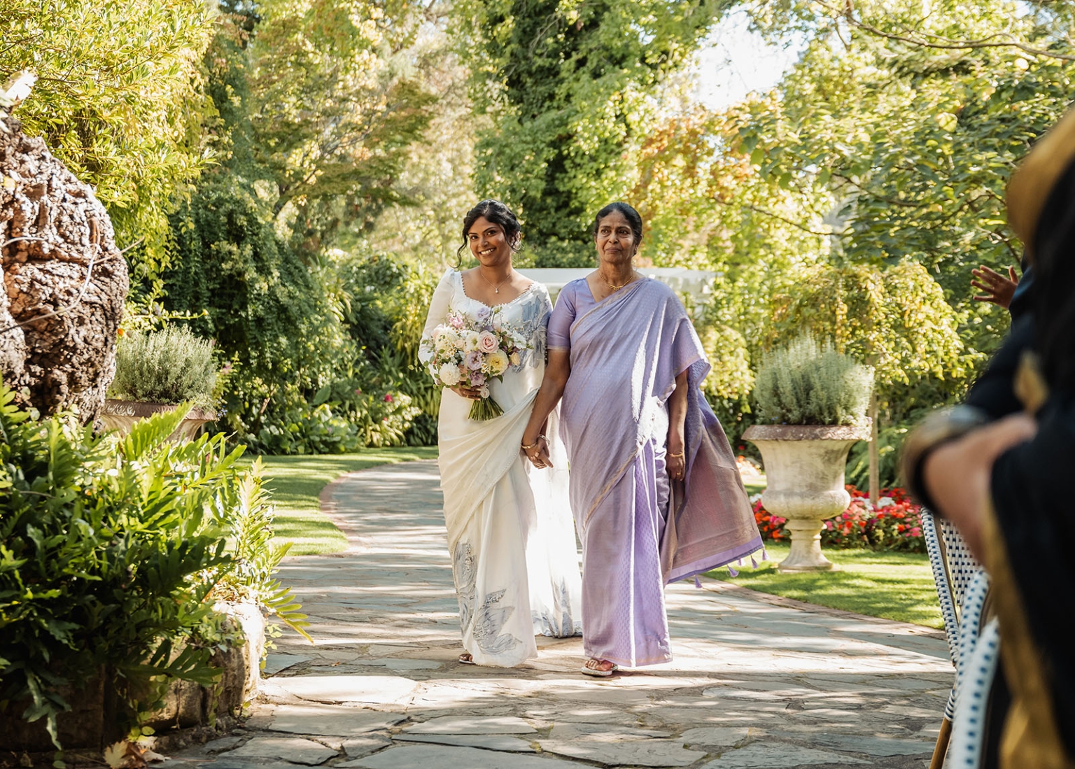 manor wedding in basket range - methmini & aravinda 132 Two women in elegant dresses walk a picturesque garden path under dappled sunlight.