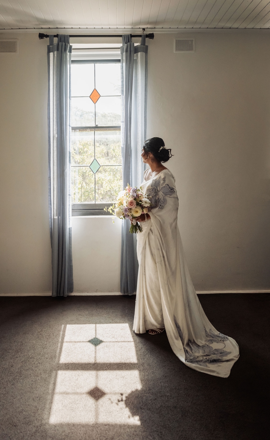 manor wedding in basket range - methmini & aravinda 119 Woman in white sari by window with flowers, in minimalistic, light-filled room.