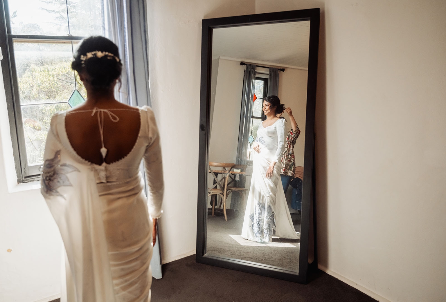 manor wedding in basket range - methmini & aravinda 114 Bride adjusts elegant wedding dress in front of mirror, highlighted by soft stained glass light.