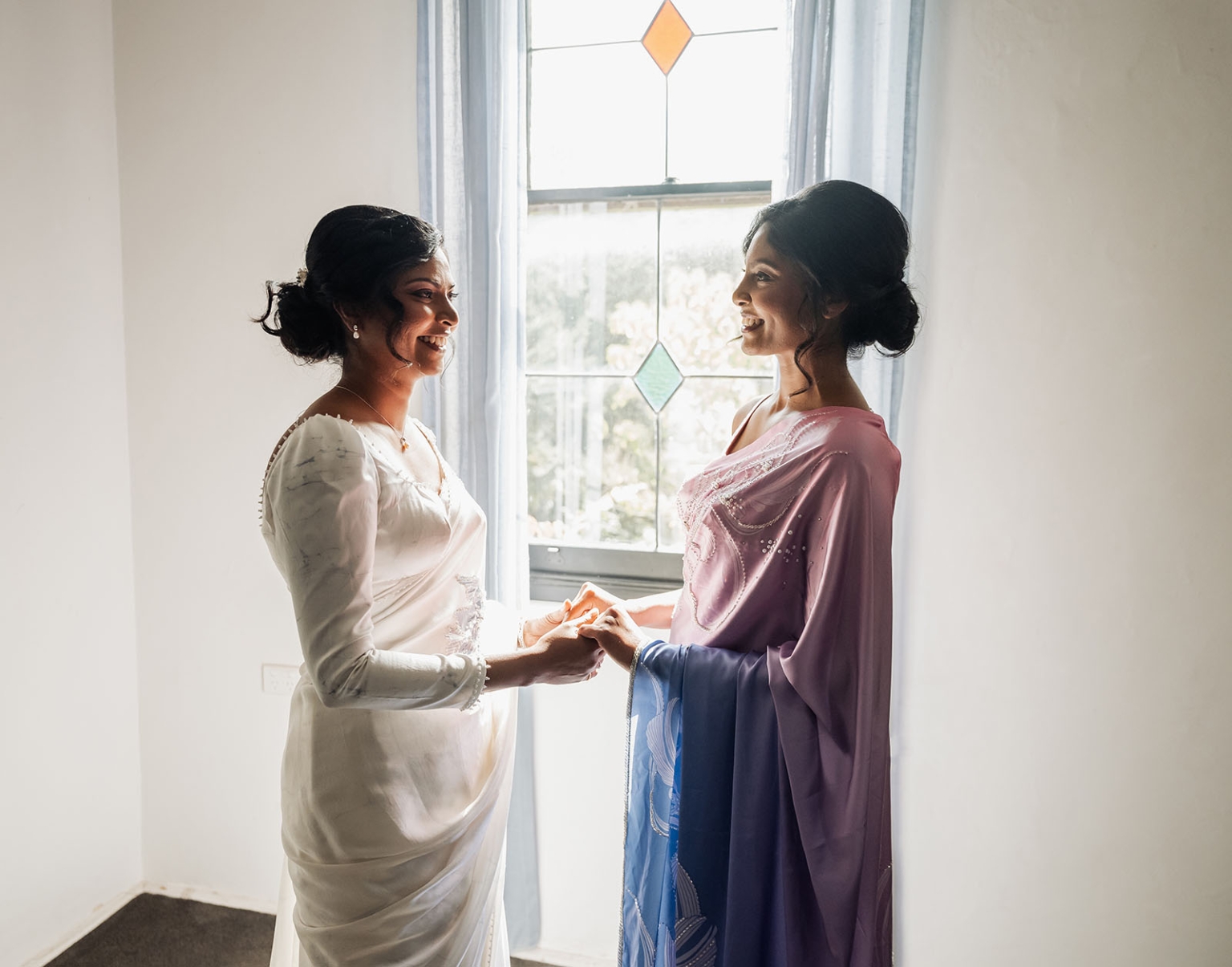 manor wedding in basket range - methmini & aravinda 118 Two women in elegant attire share a joyful moment by a sunlit window.