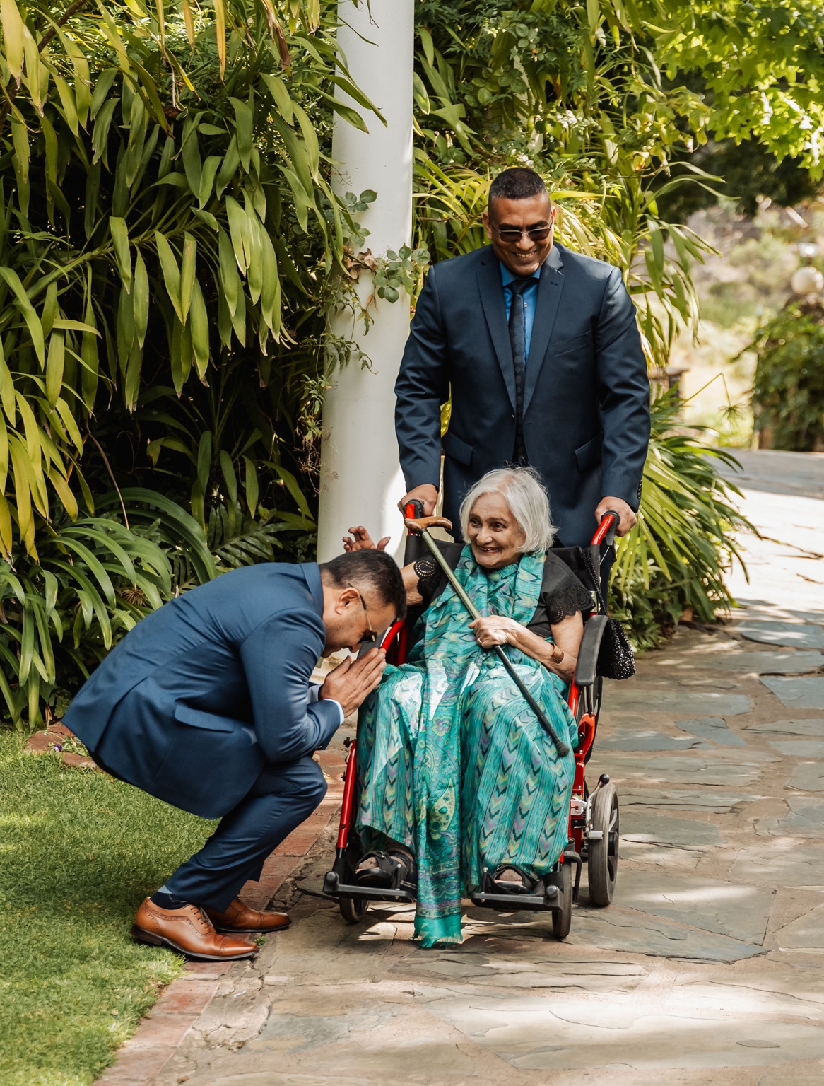 manor wedding in basket range - methmini & aravinda 130 Elderly woman in wheelchair with family in garden, expressing love and respect.