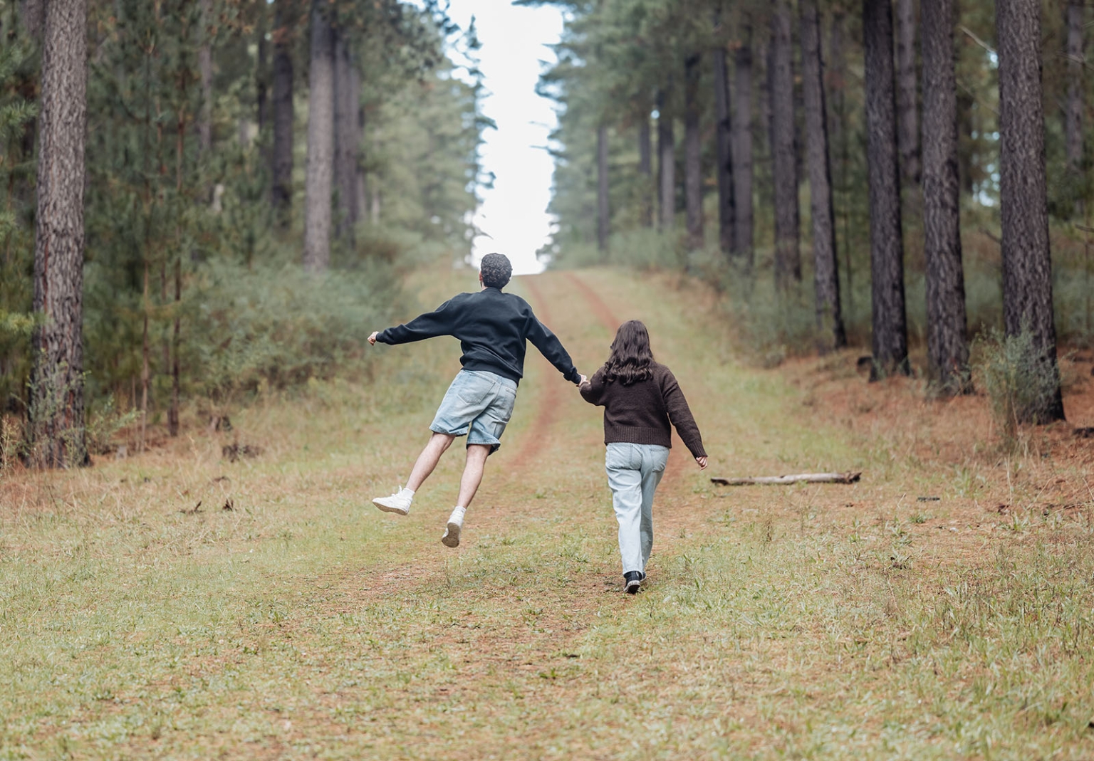 cabn at kuitpo forest proposal - tamsyn & blake 15 Couple walking and leaping through a forest path, surrounded by towering trees and lush greenery.