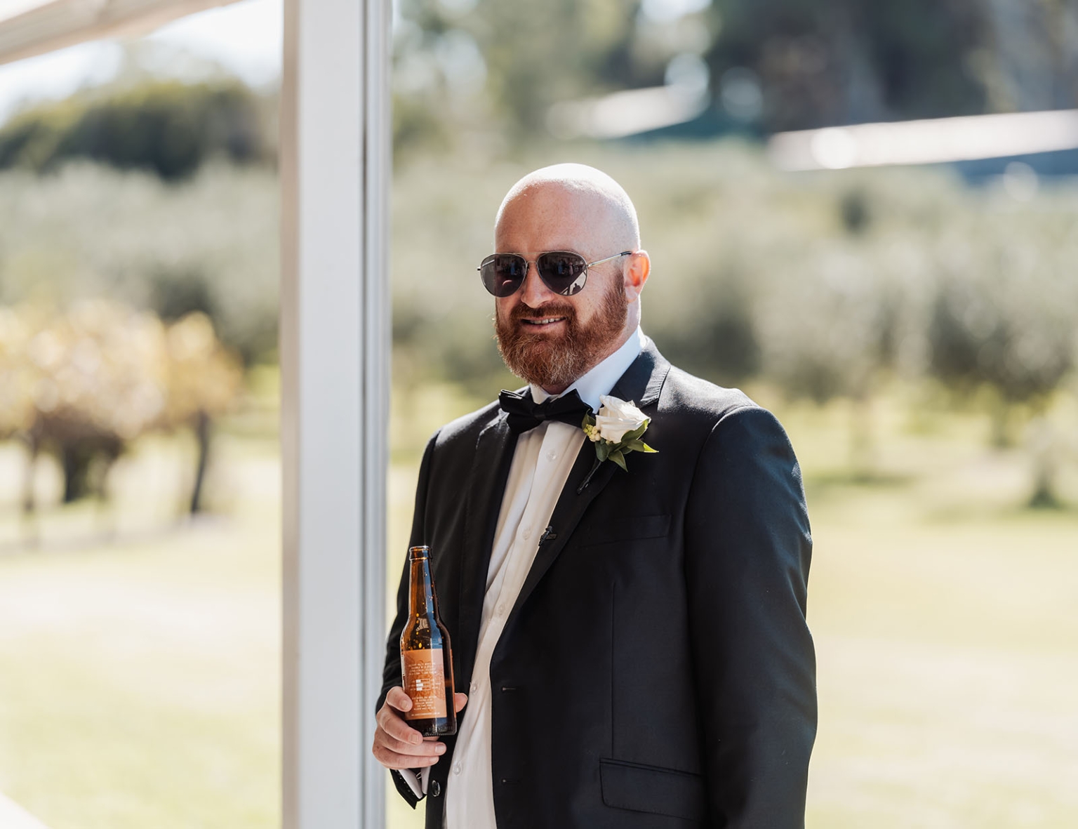 lloyd brothers wedding - dajana & tom 4 Man in tuxedo with boutonniere and sunglasses, holding bottle, outdoors at formal event.