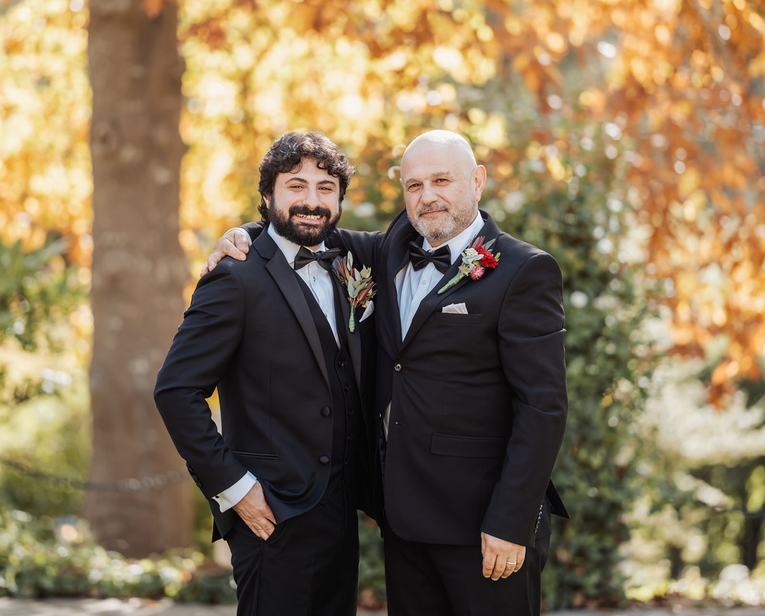 beautiful manor basket range wedding - sophie & theodore 84 Two men in tuxedos outdoors, embracing in friendship, with fall foliage background.