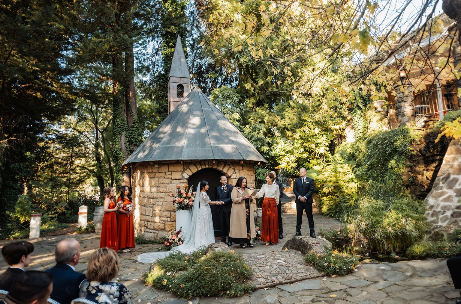 adelaide hills wedding at the manor - jo & dan 23 Outdoor garden chapel wedding ceremony with bride, groom, rustic stone chapel, and forest greenery.