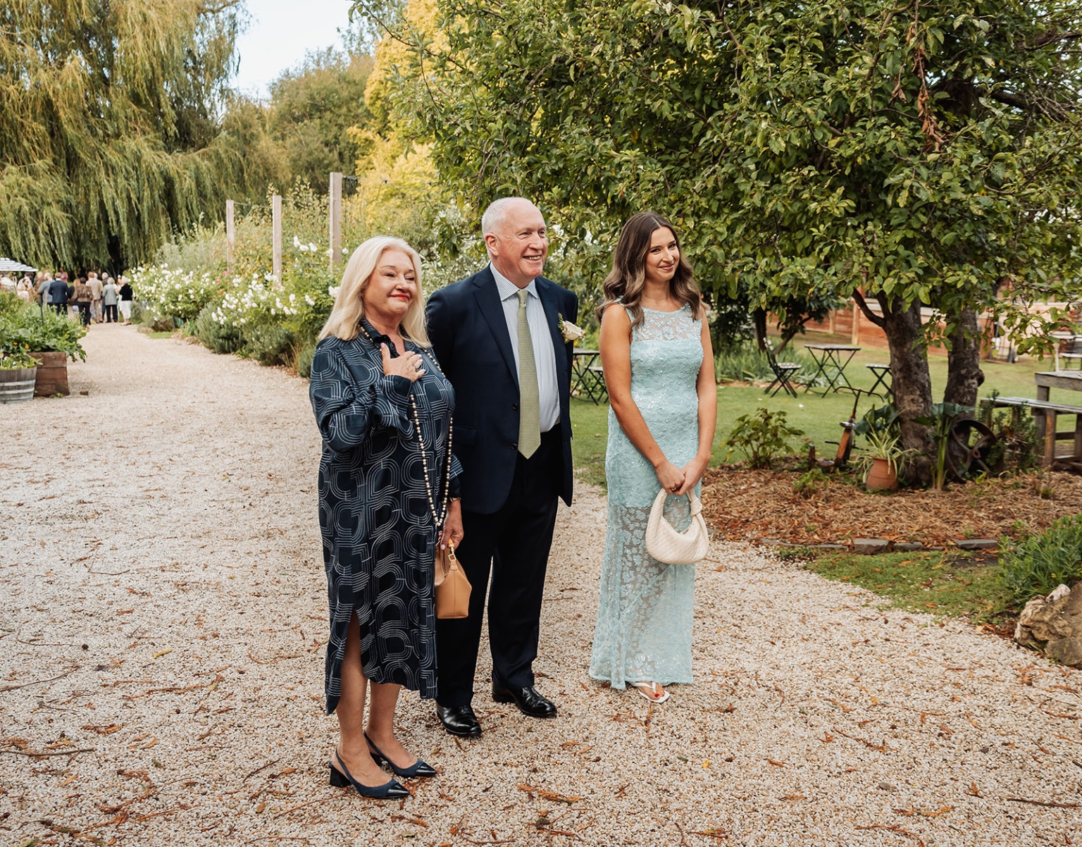 comida wedding - georgia & james 10 Three people in formal attire smiling in a lush garden setting.