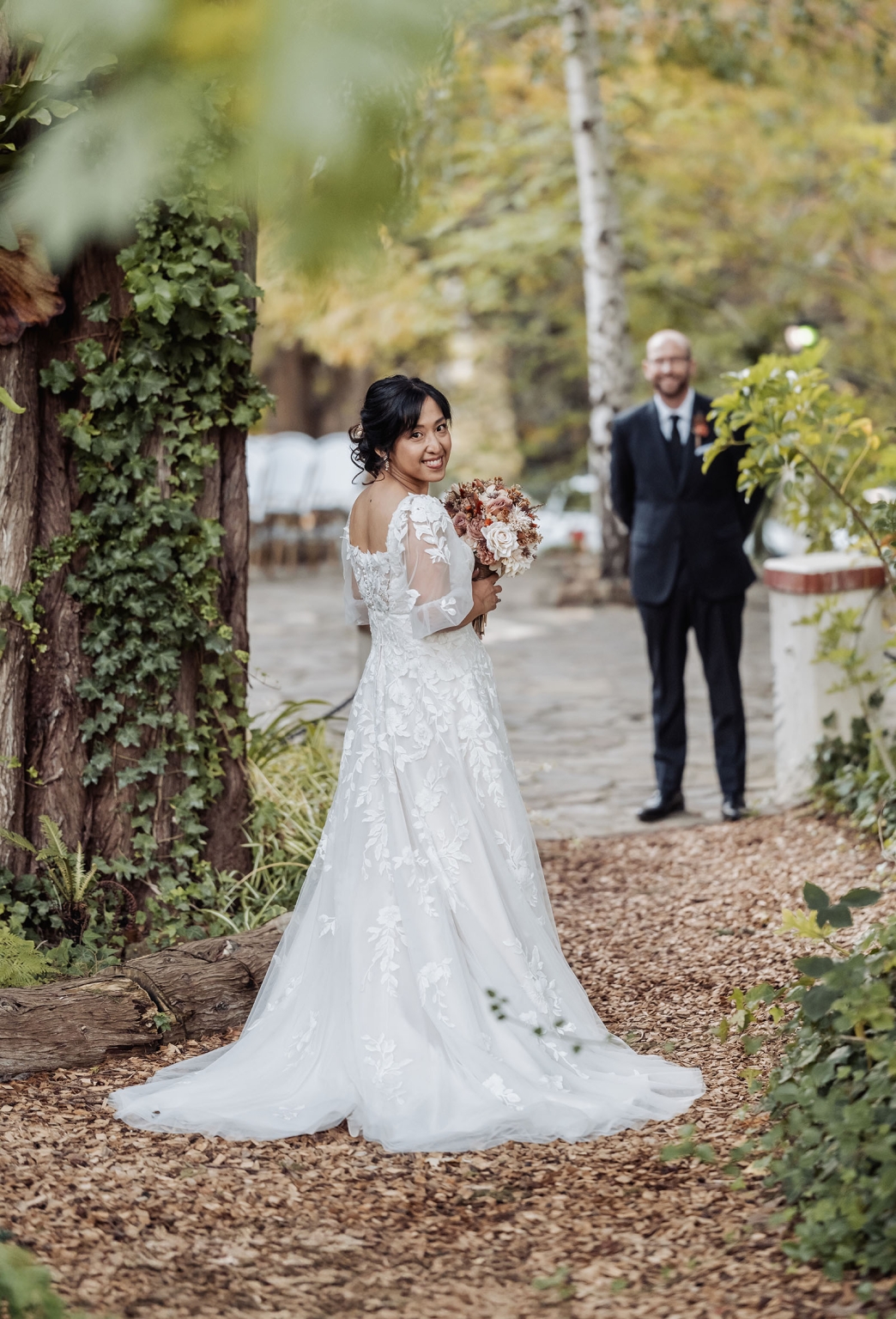 adelaide hills wedding at the manor - jo & dan 35 Garden wedding bride portrait with lace gown, bouquet, and blurred groom on wooded path.