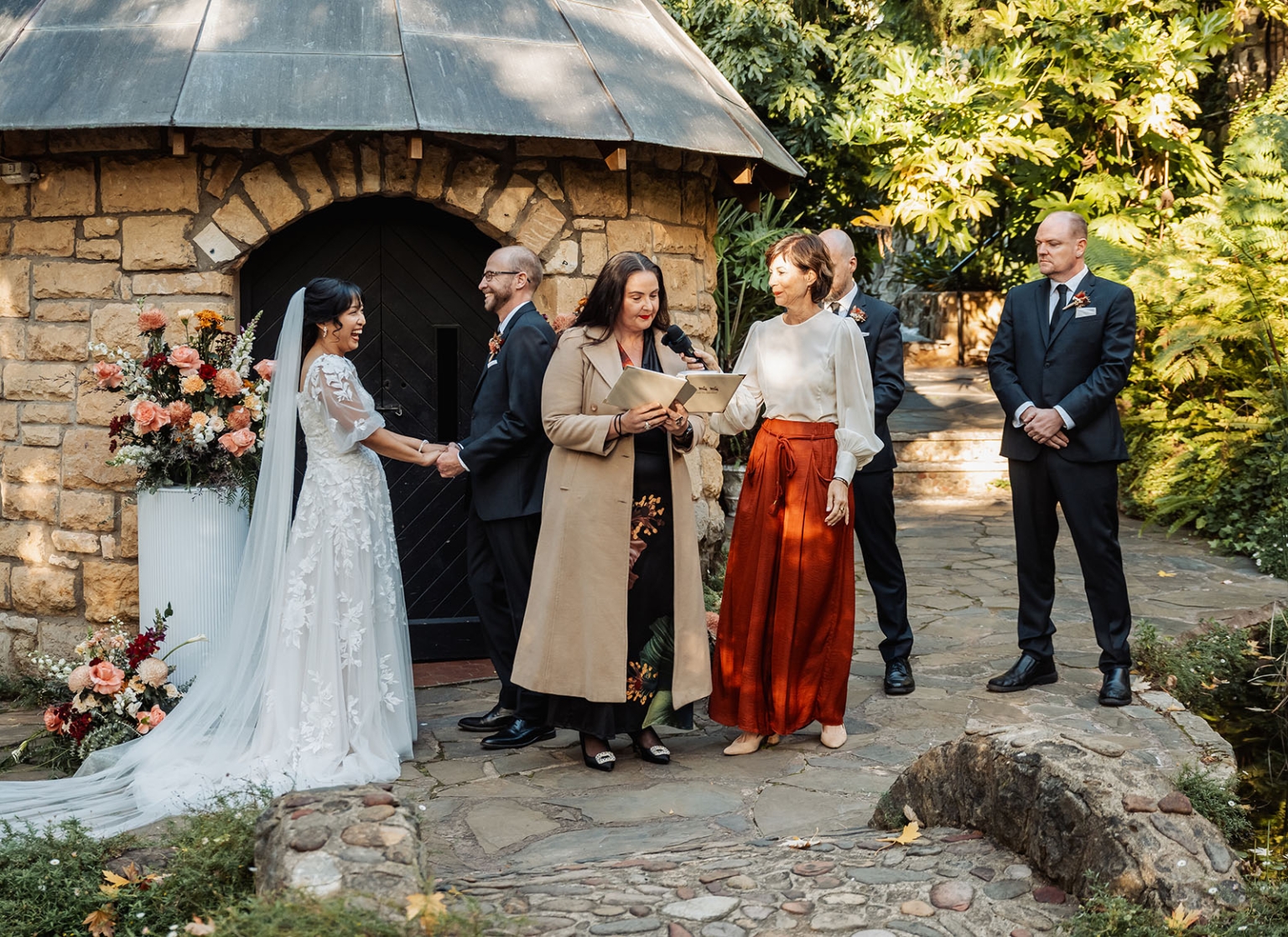 adelaide hills wedding at the manor - jo & dan 22 Garden wedding ceremony with bride, groom, officiants, and floral stone chapel backdrop.