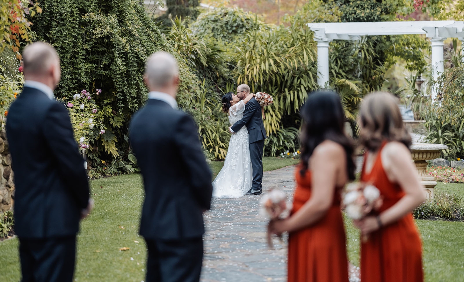 adelaide hills wedding at the manor - jo & dan 38 Bride and groom kiss in lush garden wedding ceremony with bridesmaids and groomsmen.