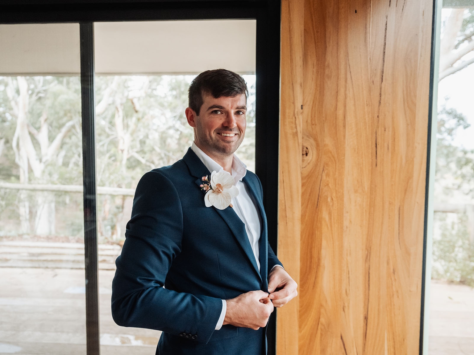 the lane vineyard wedding - caitlyn & craig 70 Man adjusts blue suit jacket with boutonniere against rustic wooden backdrop indoors.