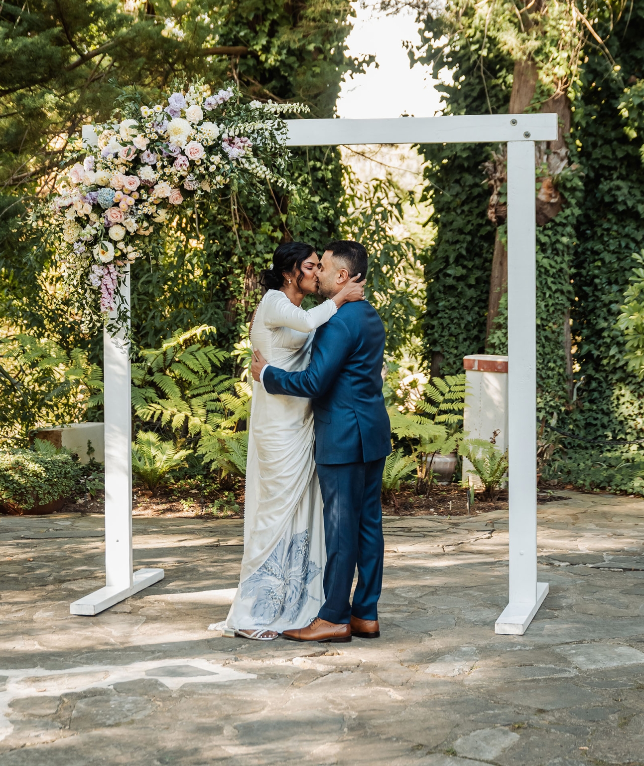 manor wedding in basket range - methmini & aravinda 142 Couple kissing under floral arch at outdoor wedding in garden setting.