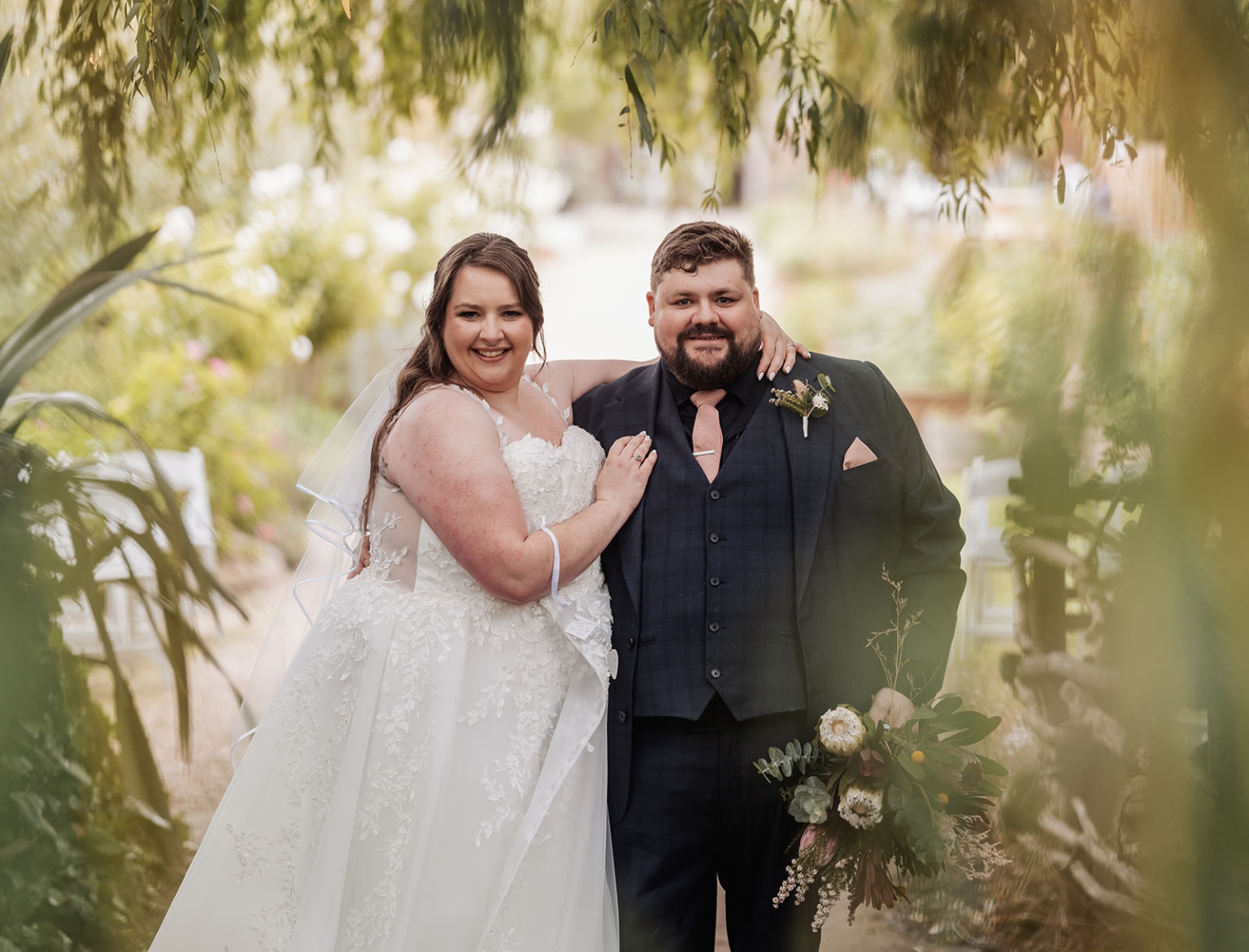 comida wedding - georgia & james 41 Bride and groom smiling in garden, wedding attire with bouquet, serene romantic setting.