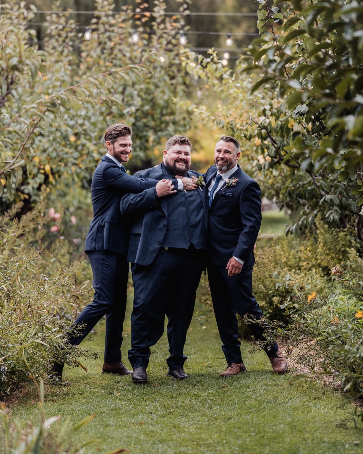 comida wedding - georgia & james 34 Three men in suits celebrate joyfully in a lush garden setting.