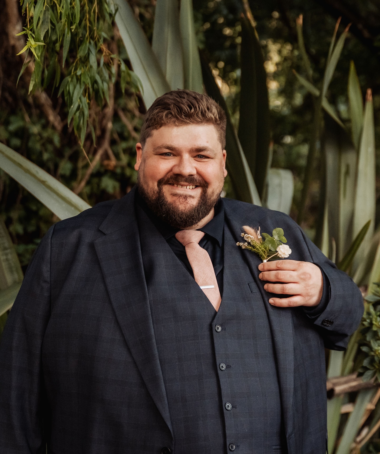 comida wedding - georgia & james 6 Man in dark checkered suit with boutonniere, smiling amidst lush greenery at a formal event.