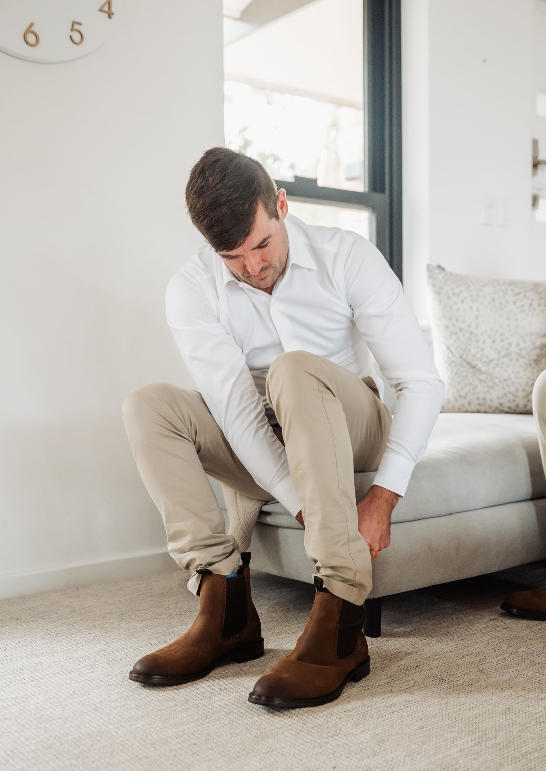 the lane vineyard wedding - caitlyn & craig 67 Man in white shirt and beige trousers putting on brown Chelsea boots in modern living room.