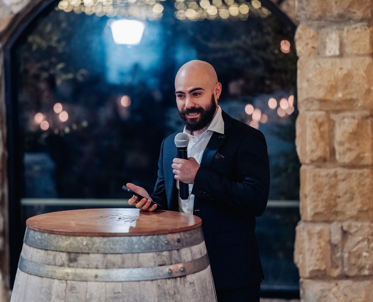 beautiful manor basket range wedding - sophie & theodore 142 Bald bearded man giving speech with microphone beside wine barrel at rustic evening event.