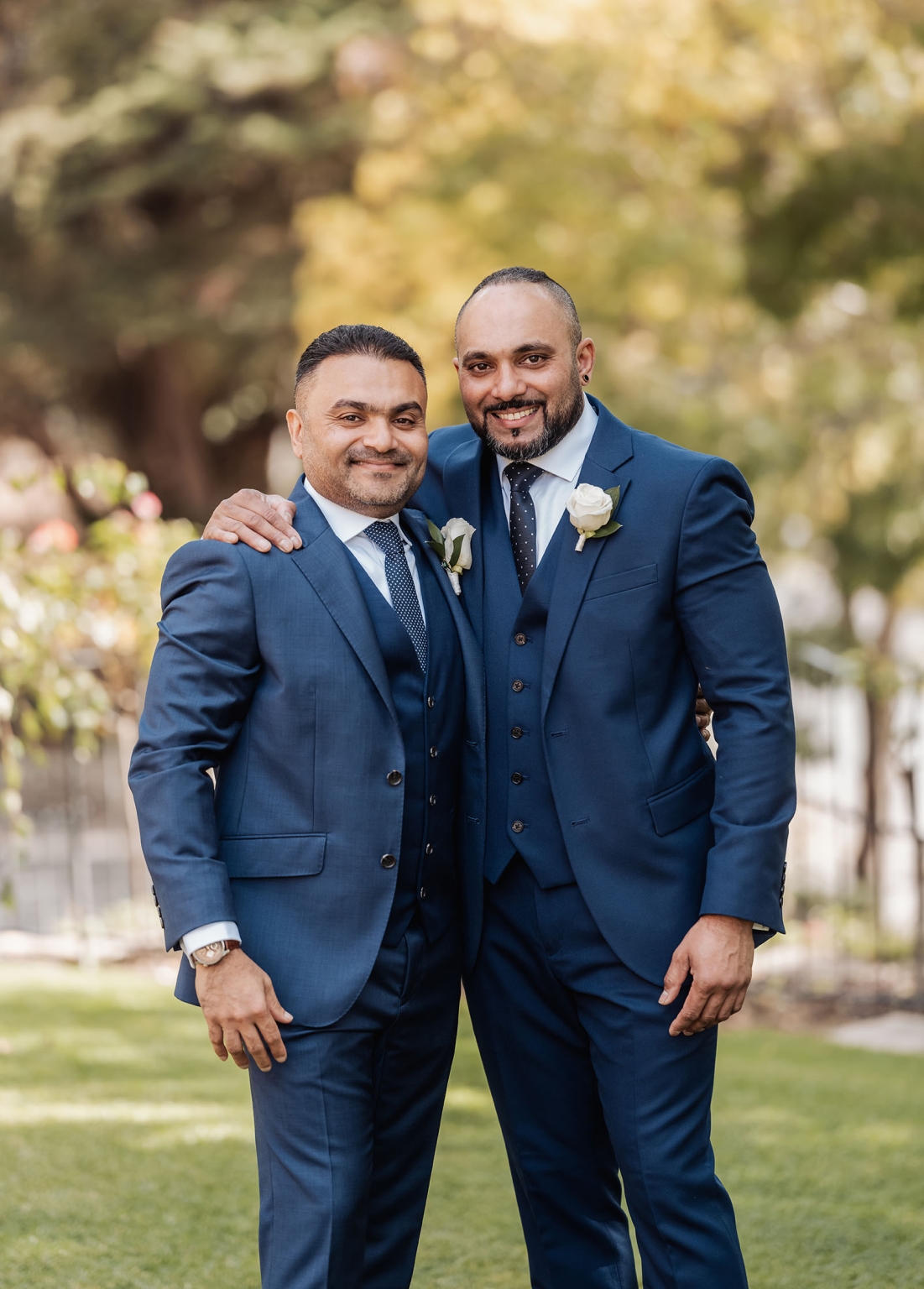 manor wedding in basket range - methmini & aravinda 103 Two men in blue suits, smiling warmly, with white roses, outdoors in a garden setting.