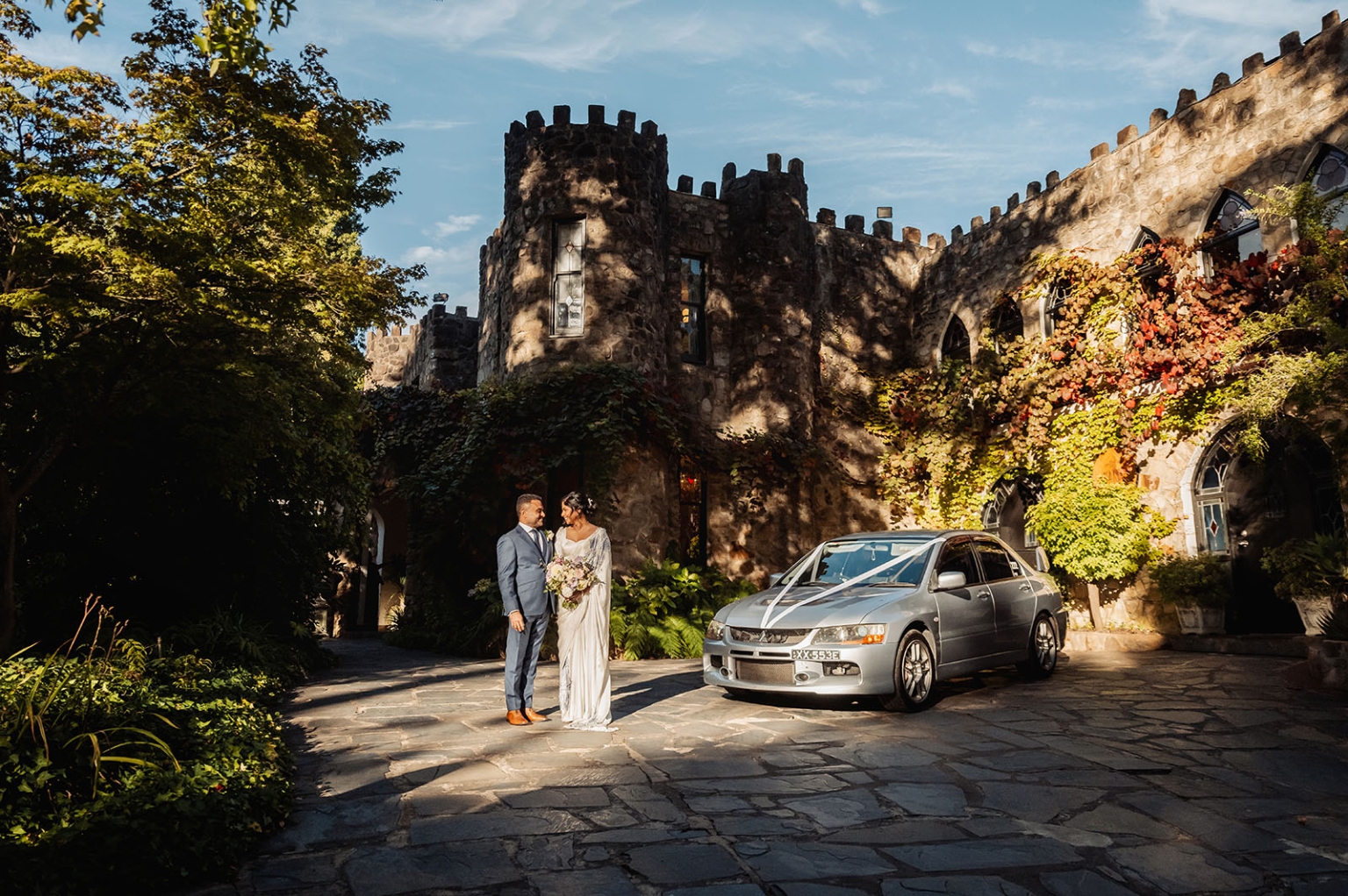 manor wedding in basket range - methmini & aravinda 152 Couple celebrates wedding in elegant castle courtyard with ivy, red foliage, and decorated car.