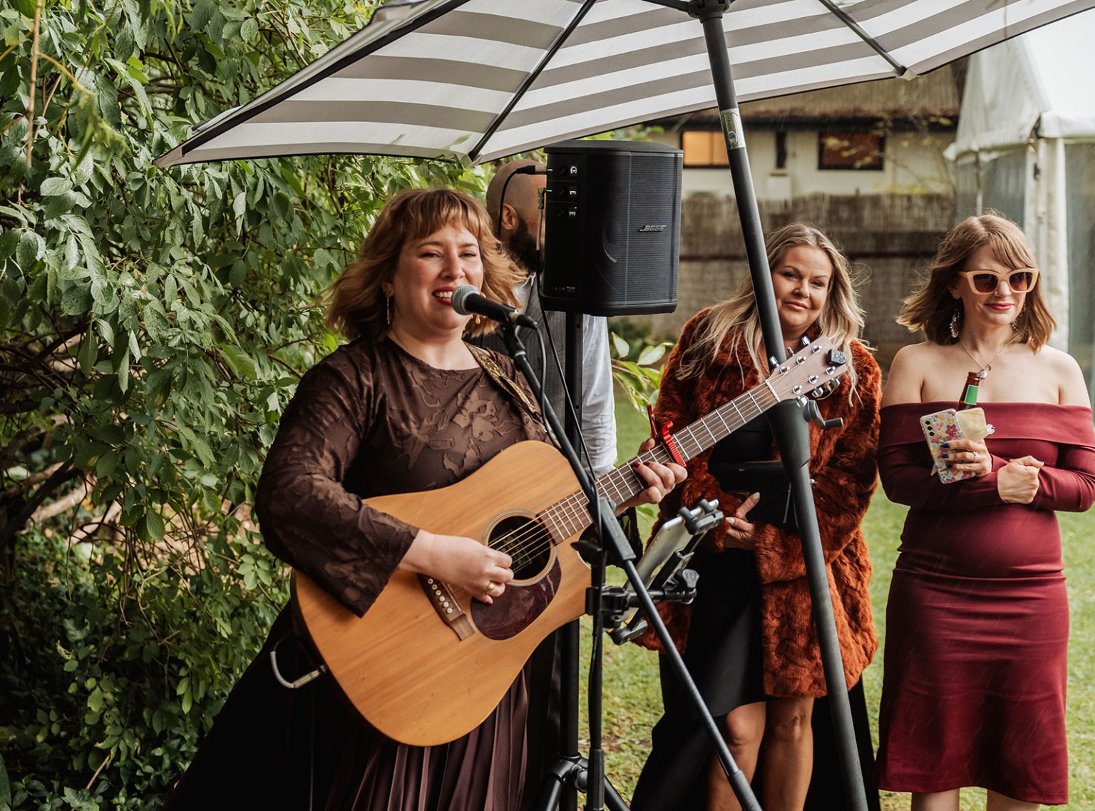 comida wedding - georgia & james 21 Female musician performing outdoors under umbrella with audience, lush greenery, and audio equipment.