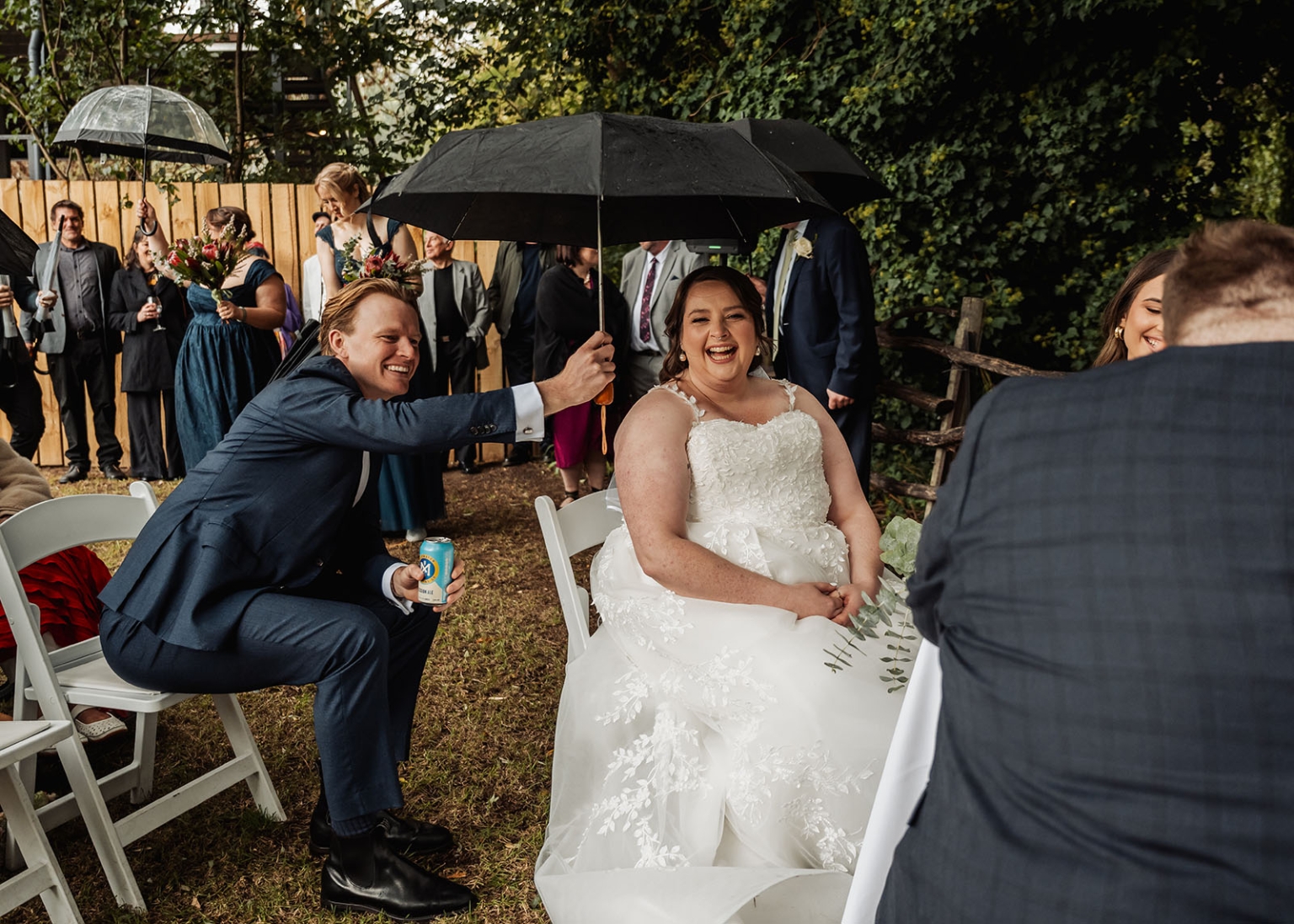 comida wedding - georgia & james 22 Bride laughing under umbrella at outdoor wedding with guests in formal attire.