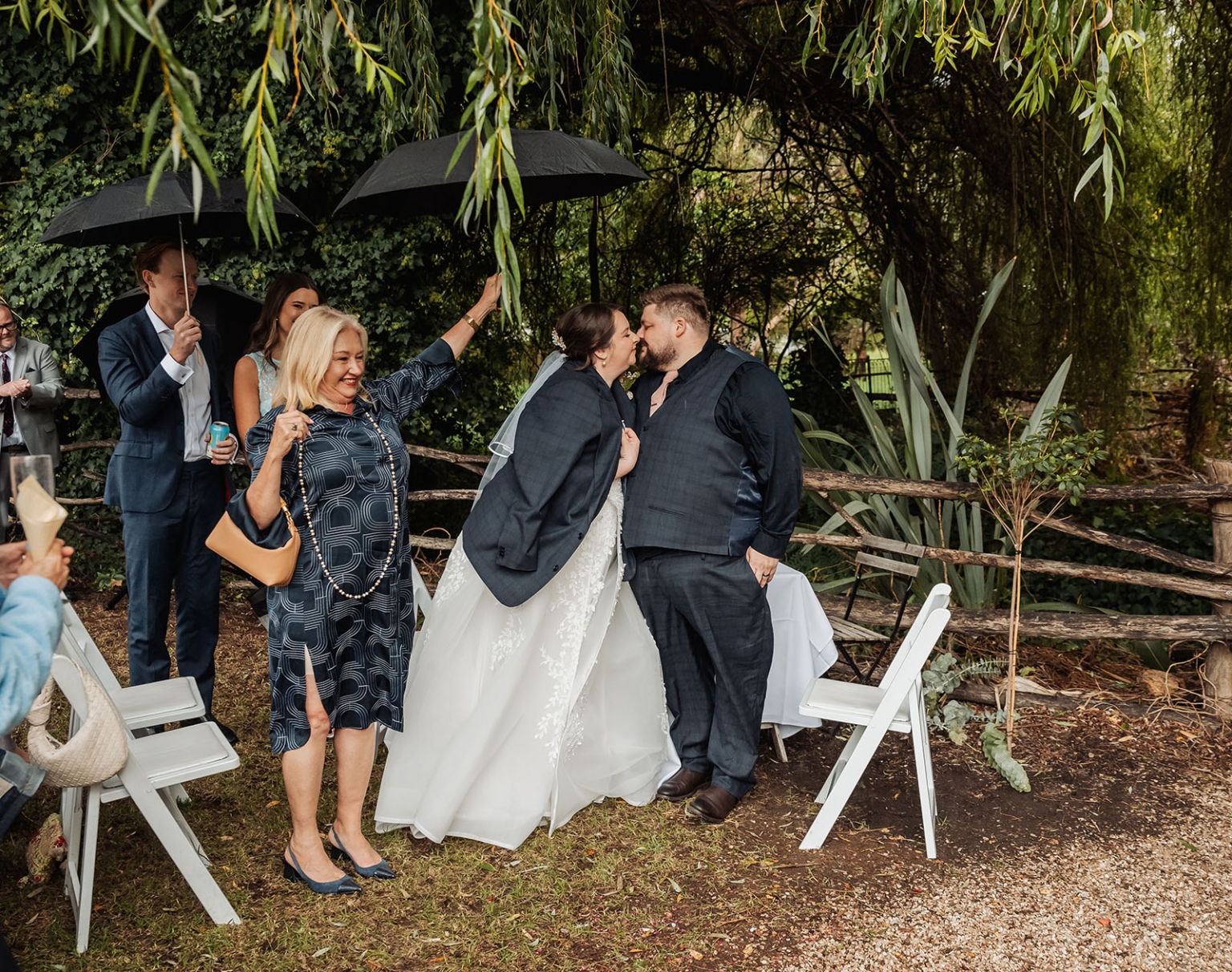 comida wedding - georgia & james 24 Bride and groom kiss under tree at outdoor wedding with guests and umbrellas.