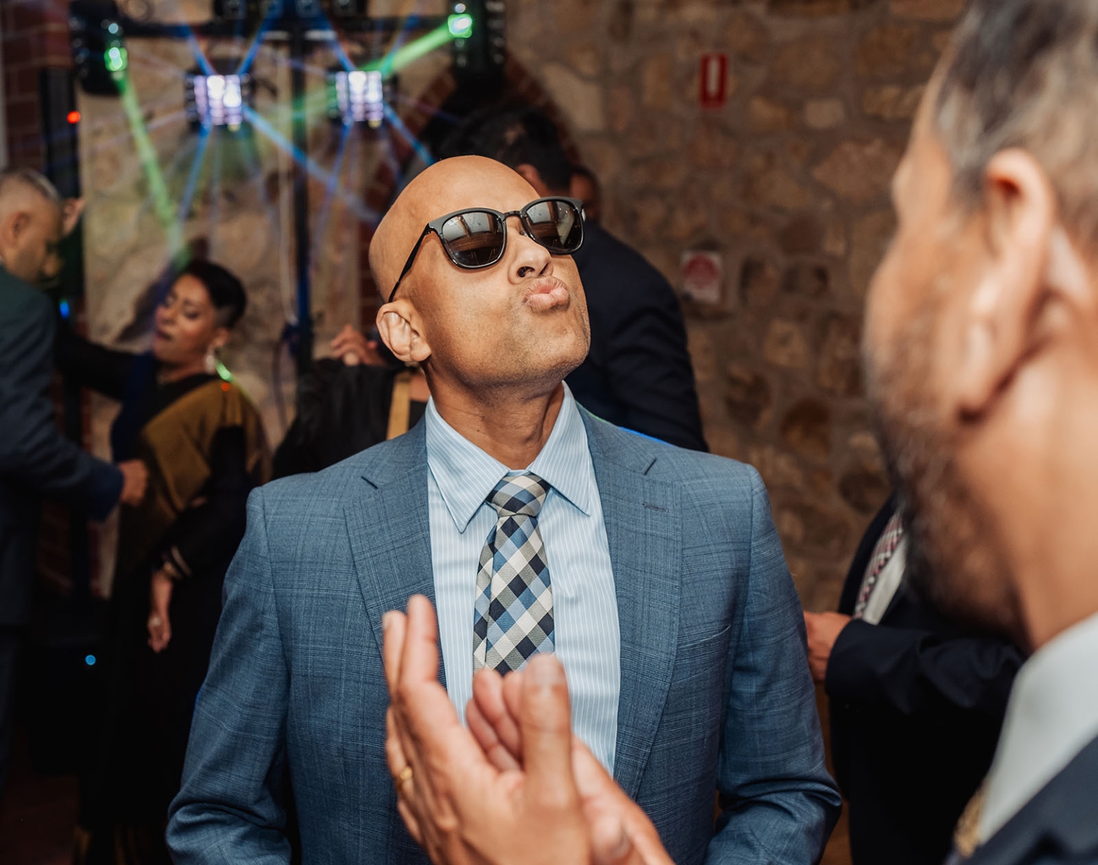 manor wedding in basket range - methmini & aravinda 171 A man in blue suit at lively party with colorful lights and stone backdrop.