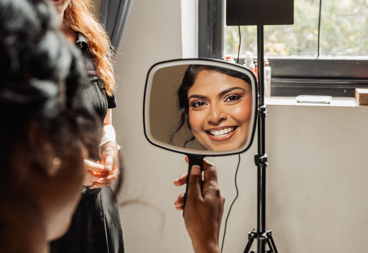 manor wedding in basket range - methmini & aravinda 110 Woman smiling in mirror, applying makeup in sunlit studio with supportive companion.