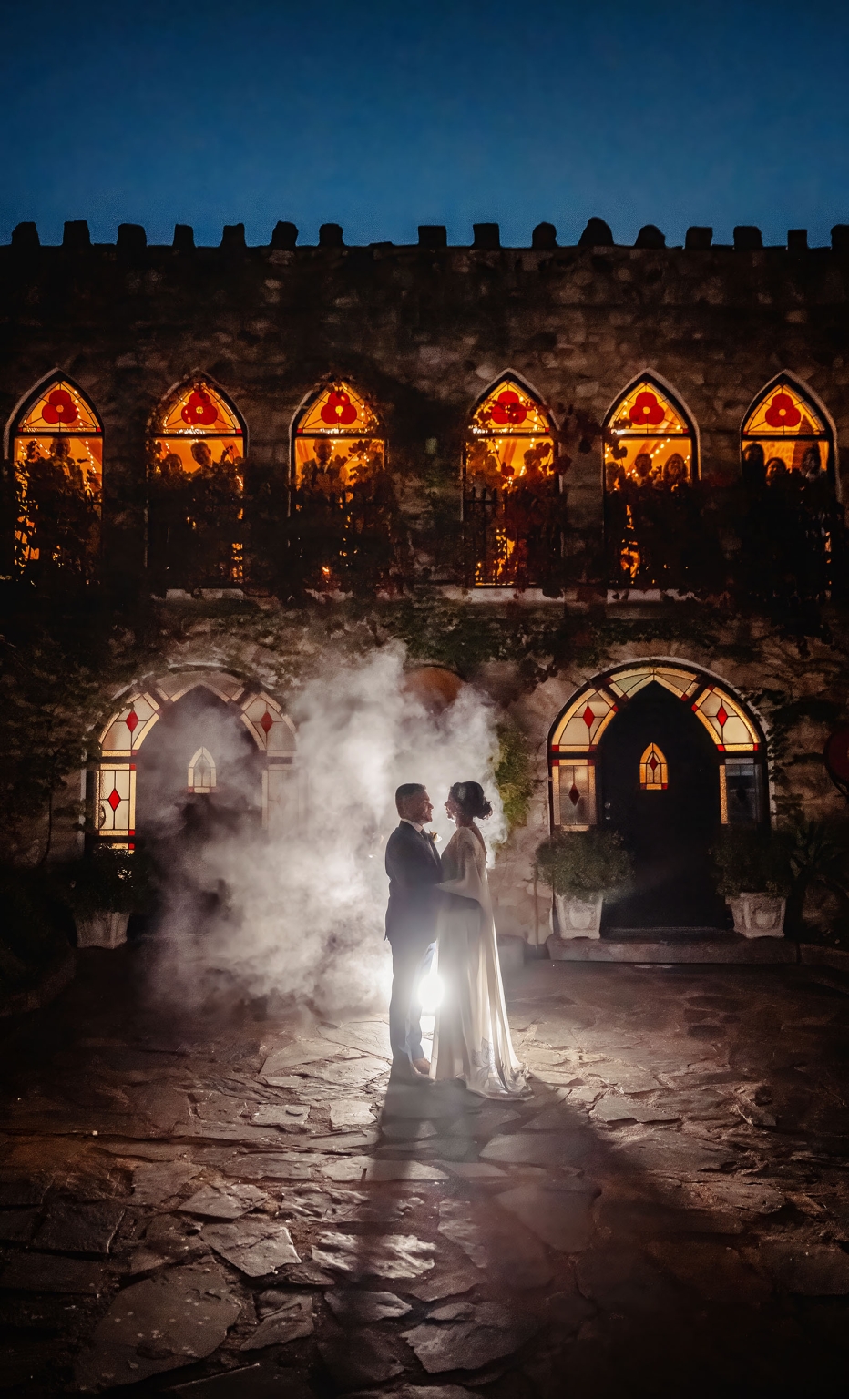 manor wedding in basket range - methmini & aravinda 163 Couple embraces before illuminated castle at night, surrounded by mist and gothic architecture.