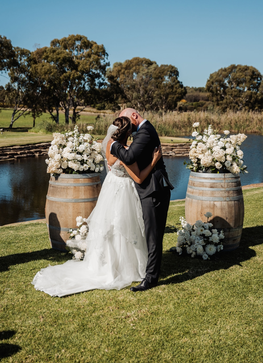 lloyd brothers wedding - dajana & tom 17 Outdoor wedding with couple kissing, elegant attire, rustic barrels, and scenic water backdrop.